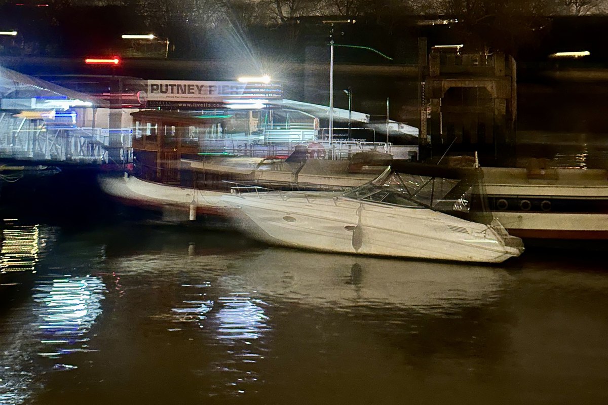 Putney Pier lit at night, a launch and a motor cruiser moored alongside, the bridge beyond and lights reflected on the water.